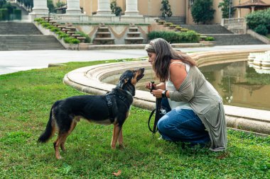 Woman kneeling in a public park, training her loyal dog with a leash, showcasing responsible pet ownership and the strong bond between humans and animals in an urban setting