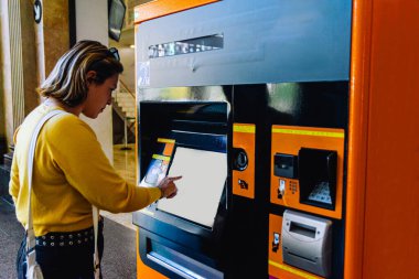 Tren istasyonunda duran genç bir kadın, otomatikleştirilmiş otomatikle toplu taşıma için bilet alıyor, seyahat planlarını bağımsız olarak yönetiyor.