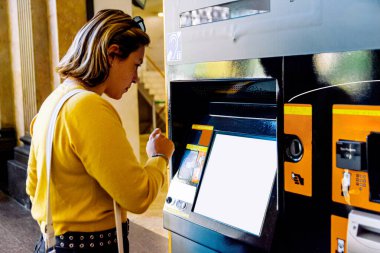 Otomatik bilet otomatında duran genç bir kadın, bir toplu taşıma istasyonunda tren bileti almak için dokunmatik ekrandan seçenekler seçiyor.