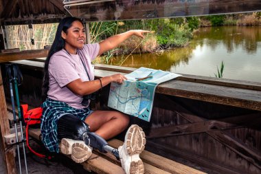 Protez bacaklı genç bir kadın kuş gözlemi altında saklanıyor, işaret ediyor, boynuna dürbün takıyor, sakin bir gölün yanında yürüyüş için güzergahını haritalıyor.