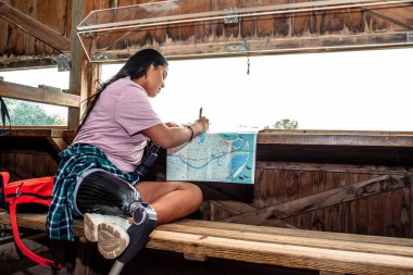 Protez bacaklı genç bir kadın tahta bir kuşun içinde kör bir şekilde oturuyor, dürbünle kuşları izliyor ve harita çalışıyor, açık hava macerasından ve aktif doğa keşiflerinden zevk alıyor.