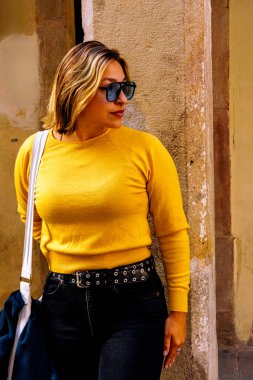 Parlak sarı kazaklı, siyah kot pantolonlu ve güneş gözlüklü bir kadın yıpranmış taş bir duvara yaslanıyor, kendinden emin, şık bir şehir profiline saldırıyor.