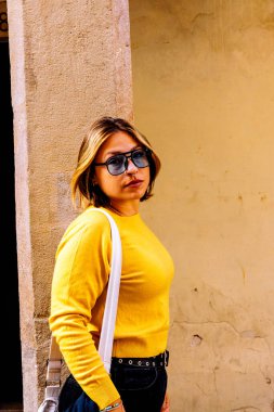 Eski bir bina duvarının yanında duran bir kadın, güneş gözlüğü, sarı bir kazak ve kemerli koyu renk kot pantolon giyerek kameraya bakıyor, kendinden emin ve şık bir şehir tavrı sergiliyor.