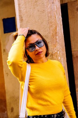 Parlak sarı kazaklı ve şık güneş gözlüklü şık bir kadın kaba dokulu bir duvara el ele poz veriyor, şehir modasını somutlaştırıyor ve rahat bir şehir hayatı sürüyor.