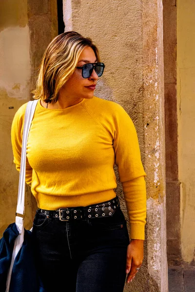 Parlak sarı kazaklı, siyah kot pantolonlu ve güneş gözlüklü bir kadın yıpranmış taş bir duvara yaslanıyor, kendinden emin, şık bir şehir profiline saldırıyor.