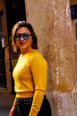 Kısa saç stili ve burun halkası takan, gülümseyen, göz kemeri takan ve canlı sarı bir kazak giyen, yaşlı, dokulu bir şehir duvarına karşı duran genç bir kadın.