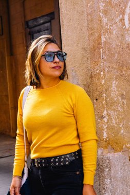 Genç bir kadın, parlak sarı bir kazak, siyah bir kot pantolon ve şık bir güneş gözlüğü giymiş, eski dokulu bir duvarın yanında durmuş düşünceli bir ifadeyle başka tarafa bakıyor.