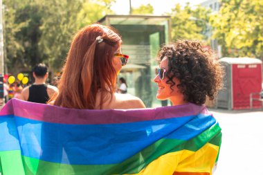 Güneşli bir açık havada gökkuşağı bayrağının altında sarılan iki neşeli kadın güneş gözlükleriyle birbirlerine gülümseyip aşkı, eşitliği ve toplumu kutluyorlar.