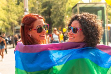 Gökkuşağı güneş gözlüğü takan ve gururlu bir bayrağa sarılı iki mutlu kadın. Güneşli bir lgbtq festivalinde gülümseyip kutlama yapıyorlar. Bu da toplumdaki sevgi, birlik ve eşitliği gösteriyor.
