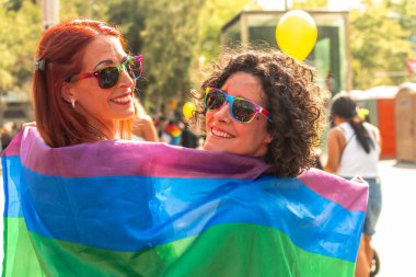 Gökkuşağı güneş gözlüklü iki kadın rengarenk bir gurur bayrağına sarılmış, açık hava gurur etkinliğinde çeşitliliği, kapsama ve neşeyi kutluyorlar, sevgi ve dayanışmayla birleştiler.
