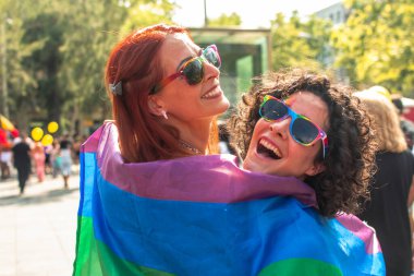 Renkli güneş gözlüğü takan ve gökkuşağı bayrağına sarılı iki neşeli kadın bir geçit töreninde neşe ve özgürlükle kutlama yaparak mutluluğu ve toplumsal dayanışmayı ifade ediyorlar.