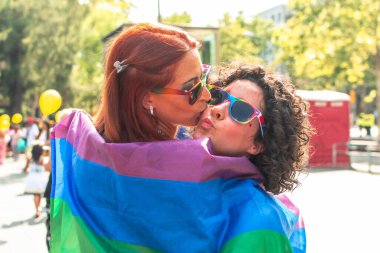 Mutlu kadınlar, gökkuşağı bayrağına sarılı ve gökkuşağı güneş gözlüğü takan, aşkı, çeşitliliği ve eşitliği kutlayan bir geçit töreninde öpüşürler.