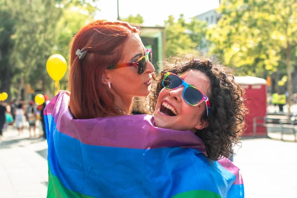 İki neşeli kadın, canlı bir gökkuşağı bayrağına sarılmış, güneş gözlüğü takmış, bir geçit töreninde aşkı ve eşitliği canlı bir atmosferle kutluyorlar.