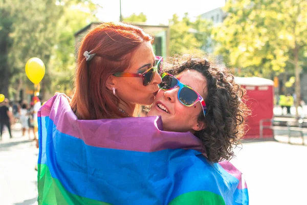 Gökkuşağı bayrağına sarılmış lezbiyen çift güneşli bir geçit töreninde neşeli bir öpüşme ve kucaklaşmayı paylaşıyor, aşkı, eşitliği, çeşitliliği ve toplumu kutluyorlar.