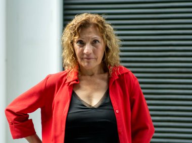 Orta yaşlı, kıvırcık saçlı, kendine güveni tam bir kadın. Açık havada kırmızı tişörtlü ve siyah bluzlu, iddialı ve kararlı bir ifade sergileyen bir kadın.