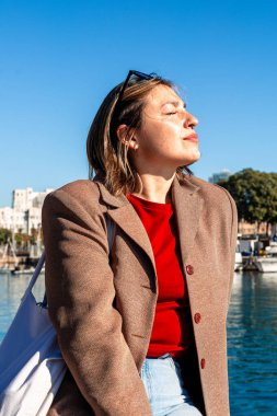 Barselona limanında dinlenen bir kadın, yüzünde güneş gözlüğü, sakin bir Akdeniz esintisi ve huzurlu, sakin bir yolculuk anı için mavi gökyüzünün tadını çıkaran bir kadın.