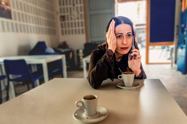 Kahve dükkanında bir masada oturan bir kadın kulağına akıllı telefon dayamış, masada iki fincan kahveyle konuşurken endişe ve stresi dile getiriyor.