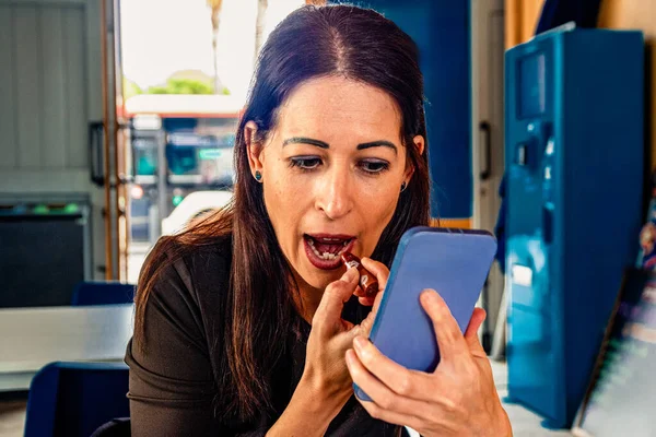Woman applying red lipstick in a casual setting while looking at her smartphone for a mirror, focusing on her appearance and personal grooming in a modern, on the go lifestyle