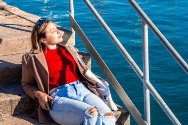 Metal korkulukların yanında güneşlenen, güneşin sıcaklığını hisseden ve limanın ışıldayan mavi suyunun keyfini çıkaran günlük giysiler içinde bir kadın.