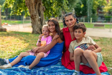 Anne ve üç çocuğu parkta bir battaniyeye oturmuş, sıcak yaz güneşini emerken ve doğada birlikte olmanın mutluluğunu kucaklarken keyifli bir pikniğin tadını çıkarıyorlar.