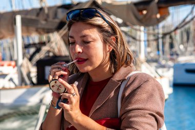 Dışarıda duran bir kadın, küçük bir aynayla dudaklarına dudak parlatıcısı sürüyor. Güzellik rutinine ve rıhtımdaki yaşam tarzına odaklanıyor.