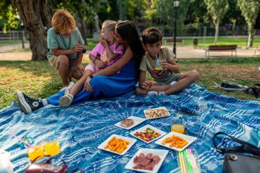 Anne ve çocuklar parkta bir battaniyenin üzerinde keyifli bir pikniğin tadını çıkarıyorlar, güzel doğal ortamlarda birlikte neşeli anılar yaratırken atıştırmalıkların ve içkilerin tadını çıkarıyorlar.