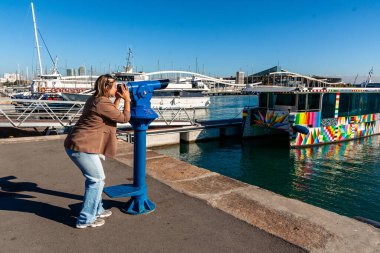 Barselona limanında güneşli bir günde kalabalık marina ve renkli katamaran teknelerini görmek için bozuk parayla çalışan bir teleskop kullanan bir kadın. İspanya 'da.