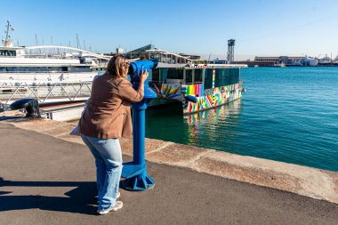 Limanın yanında duran bir kadın, liman faaliyetlerini ve yatları gözlemlemek için bozuk para kullanan bir teleskop kullanıyor, Barselona, İspanya 'da güneşli bir gezi ve turizm gününün tadını çıkarıyor.