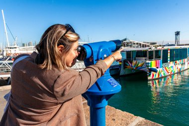 Bir kadın iskelede durmuş, Barselona Limanı 'nın manzarasından zevk alıyor, bozuk parayla çalışan bir teleskop kullanıyor ve parlak, güneşli bir günde renkli tekneleri ve manzaralı limanı işaret ediyor.