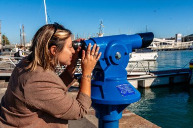 Açık güneşli bir günde mavi turist teleskopuyla ufka odaklanan, Barselona limanını, botları ve Akdeniz kıyılarını izleyen ve manzaranın tadını çıkaran bir kadın.