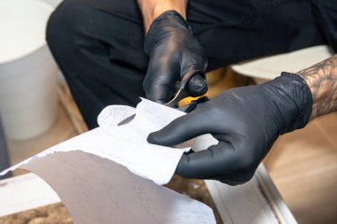 Siyah koruyucu eldivenler giyen dövme sanatçısı, küçük makasla bir şablon tasarımını dikkatlice kesiyor, stüdyoda yapılacak bir vücut sanatı seansı için özel bir kalıp hazırlıyor.