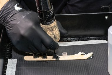 Siyah eldivenler giyen sanatçı dövme ekipmanları hazırlıyor, makinenin iğnesini küçük bir tahta çubukta siyah bir mürekkebe batırıyor, dövmeye hazır.