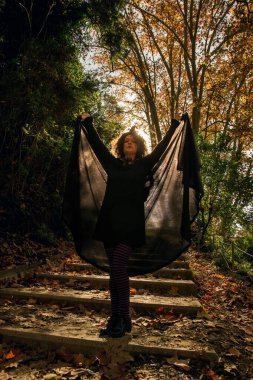 Cadılar bayramı ya da karnaval için cadı gibi giyinmiş bir kadın, kollarını kaldırmış beton merdivenlerde kara bir pelerin tutuyor orman parkında düşen sonbahar yapraklarıyla kaplı.