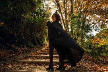 Sivri uçlu şapkalı ve pelerinli bir kadın düşen sonbahar yapraklarıyla kaplı beton merdivenleri tırmanıyor Cadılar Bayramı 'nı, karnavalını ya da ormandaki fantazi temalarını somutlaştırıyor.