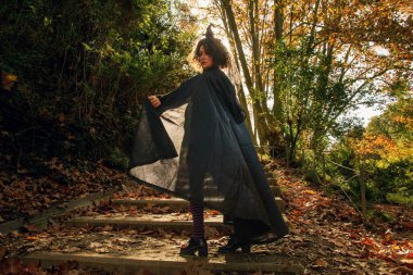 Cadı kostümü giymiş, sivri uçlu şapkalı ve uzun siyah pelerinli bir kadın. Güneşli bir sonbahar ormanında yapraklarla kaplı taş basamaklarda yürüyor. Sihir ve fantema tasvir ediyor.