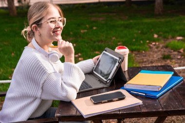 Genç kız öğrenci kampüsteki ahşap bir masada oturuyor, elinde bir kalem tutarken düşünceli bir şekilde gülümsüyor, dışarıda çalışmak için dizüstü bilgisayarla, tabletle, telefonla, kitaplarla ve kulaklıkla çevrili.
