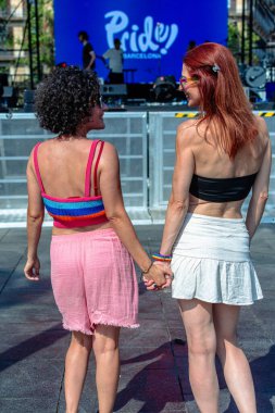 İki kadın el ele tutuşuyor ve arkadan gülümsüyor. Güneşli bir günde arka planda görünen bir sahne ile halka açık bir lgbtq + festivalinde gurur aksesuarları sergiliyorlar.