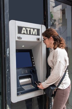 Otomatik veznedar makinesinin başında duran bir kadın, elinde para transferi için akıllı bir telefon tutuyor, bankadan bağlantısız bir şekilde para çekiyor.
