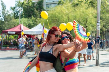 Güneş gözlüğü takan ve renkli kıyafetler giyen iki neşeli kadın kucaklaşıyor ve el ele tutuşuyor, coşkulu bir lgbtq artı gurur festivaline katılıyor, balonlar ve katılımcılarla çevrili