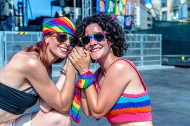 Gökkuşağı aksesuarlı iki gülümseyen kadın açık hava onur geçidinde el ele tutuşuyorlar, aşkı, çeşitliliği ve dayanışmayı neşe, destek ve renkli bir ifadeyle kutluyorlar.