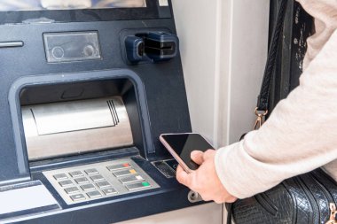 Otomatik veznedar makinesinin yanında elinde akıllı bir telefon bulunan kişiler, temassız bir ödeme veya para çekme için, modern bankacılık, finans teknolojisi ve dijital cüzdan kolaylığını sembolize ediyor.