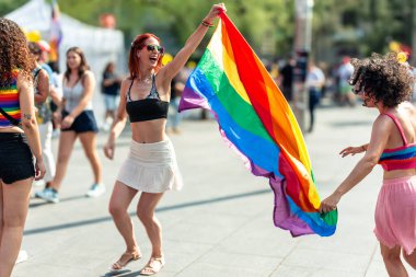 Neşeli bir kadın arkadaşlarıyla dışarıda kutlama yaparken ve yaz ayinlerinde özgürlük ve çeşitliliği ifade ederken gururlu bir gökkuşağı bayrağı sallıyor.