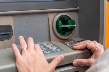 Kişinin elleri ATM ile etkileşime giriyor, kredi kartı takıyor ve banka hesaplarını yönetmek ve güvenli bir işlemi tamamlamak için tuş takımına bir pim takıyor.