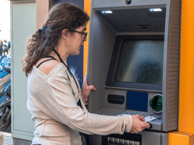 Otomatik veznedar makinesini kullanan bir kadın - ATM - kentsel bir ortamda, kredi kartını yuvaya sokarak, kişisel finansmanı yöneterek ve nakit çekerek finansal işlem gerçekleştiriyor.