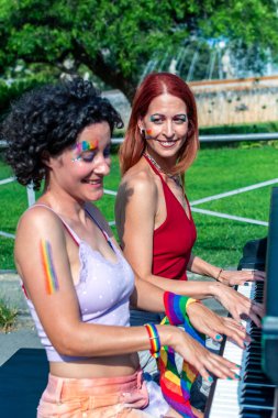 Gökkuşağı makyajlı ve aksesuarlı iki gülümseyen kadın açık havada piyano çalıyor, çeşitliliği, eşitliği ve gururu, yemyeşil bir güneşli parkta müzik eşliğinde kutluyorlar.