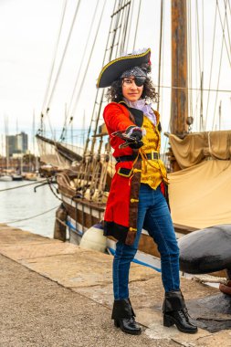 Kendine güvenen, korsan kostümü giyen, göz bandı ve şapkası olan genç bir kadın, limanda eski bir yelkenli gemisiyle dikilirken elinde bir kılıç tutuyor.