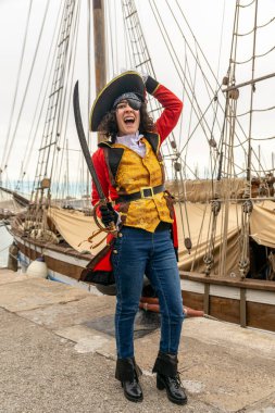 İskelede korsan kostümü giymiş bir kadın, limandaki şenlik şenliğinde tarihi bir geminin yanında göz bandı, şapka ve kılıçla gülüyor ve poz veriyor.