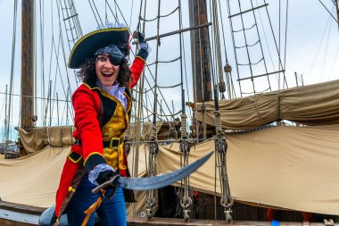 Neşeli korsan kadın, tarihi bir yelkenli gemisinin güvertesinde kırmızı bir kostüm giymiş, elinde bir kılıç tutan ve kameraya hevesle poz veren bir kadın.