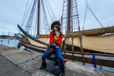 Korsan gibi giyinmiş, göz bandı ve kılıçla gülümseyen kişi, limanda geniş bir yelkenli teknesinin yanında, bulutlu bir gökyüzünün altında, karnaval sezonunu kutluyor.