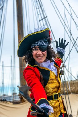 Tricorne şapkası, göz bandı ve kırmızı ceketi olan korsan kostümlü bir kadın güvertede bir kılıç sallıyor, gülümsüyor ve denizde canlı bir karnaval festivalinde kutlama yapıyor.